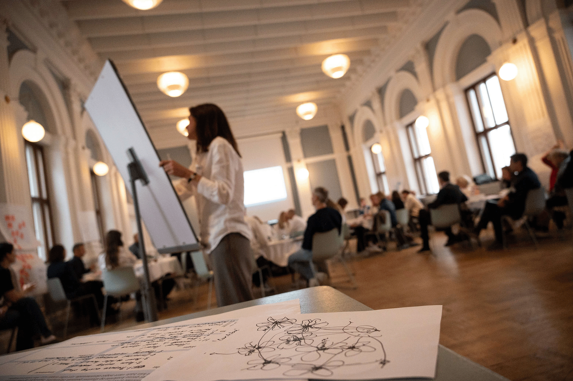Facilitator leading a workshop in a bright, high-ceilinged room with participants seated at round tables, engaged in group discussions.