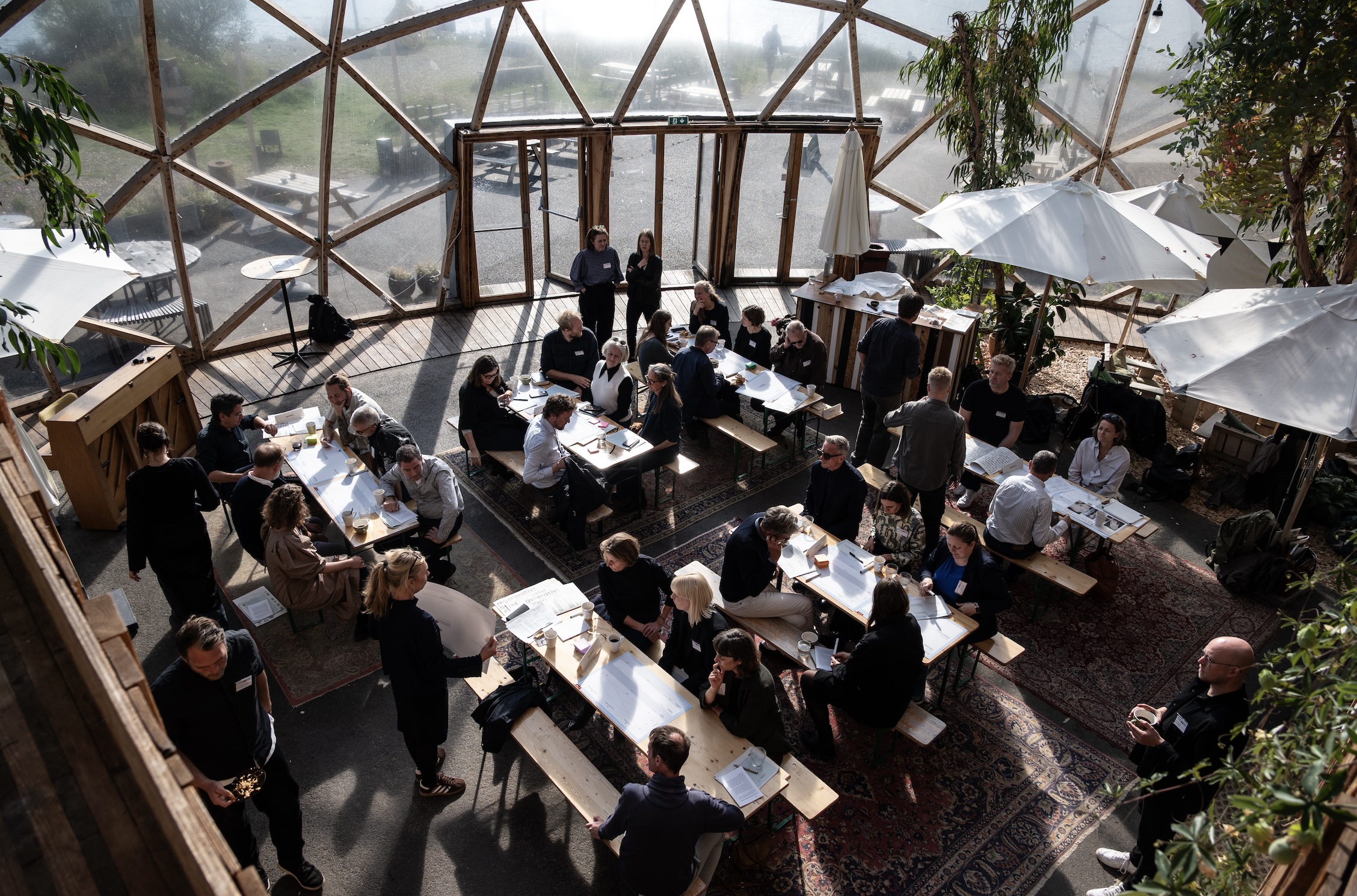 Gruppe af mennesker samlet ved lange træborde i en lys glaskuppel under et workshop- eller netværksarrangement